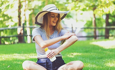 Young woman wearing a sun hat, sitting on the grass applying sunscreen