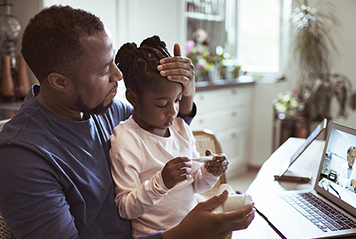 Father feeling forehead of sick daughter, speaking with healthcare provider on video call