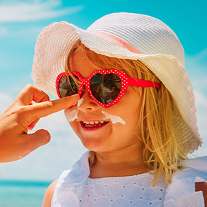  Young girl in a hat and sunglasses having sunscreen applied to her face