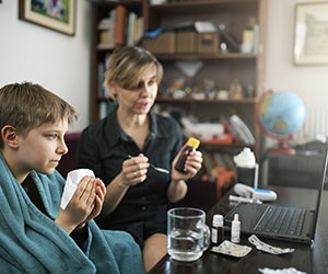 Sick child holding tissue to face; adult woman on videochat with healthcare provider